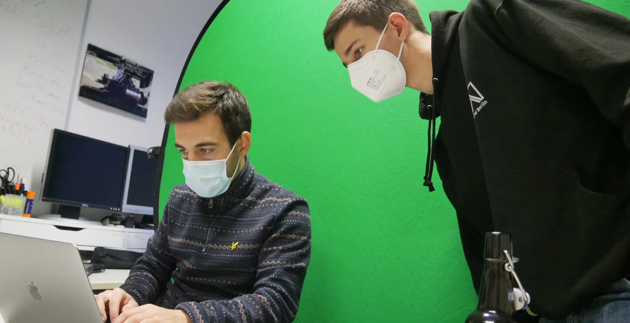 Zwei CoderDojo-Mitglieder bereiten ein Workshop-Video vor. Im Hintergrund befindet sich ein Greenscreen. Philipp (links) arbeitet am Laptop, während Luis (rechts) ihm über die Schulter sieht.